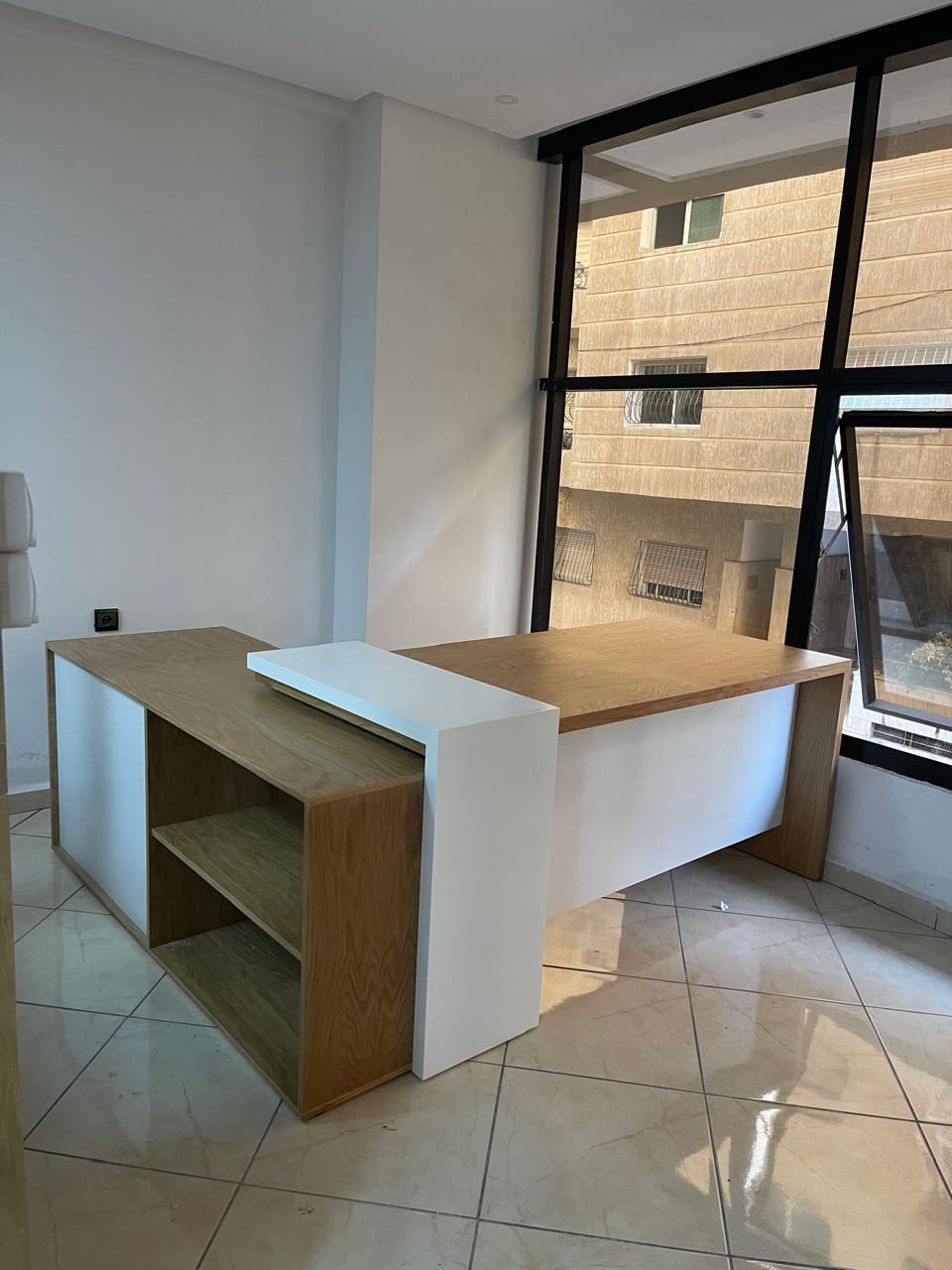 Bureau d'angle blanc et bois clair avec retour et niches de rangement ouvertes, devant une baie vitrée.