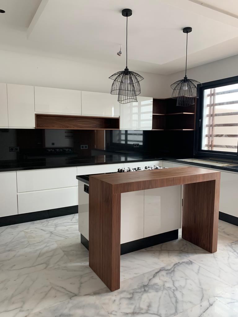 Cuisine sur mesure à façades blanches sans poignées, îlot central en bois foncé, crédence en verre noir et suspensions métalliques noires.