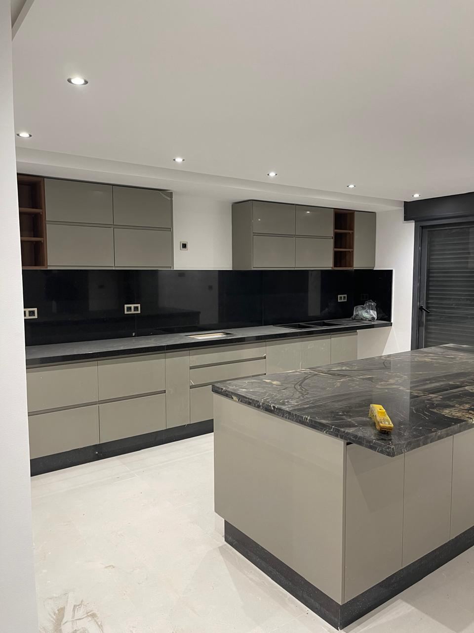 Cuisine en longueur aux façades gris taupe, crédence en verre noir, et îlot central au plan de travail en granit sombre.
