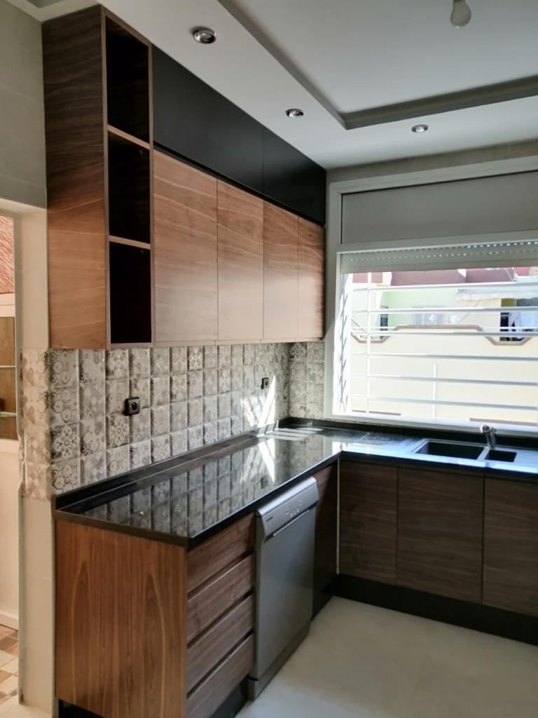 Cuisine en L mêlant façades en bois foncé et caissons noirs, crédence en pierre appareillée, évier deux bacs et plan de travail noir.