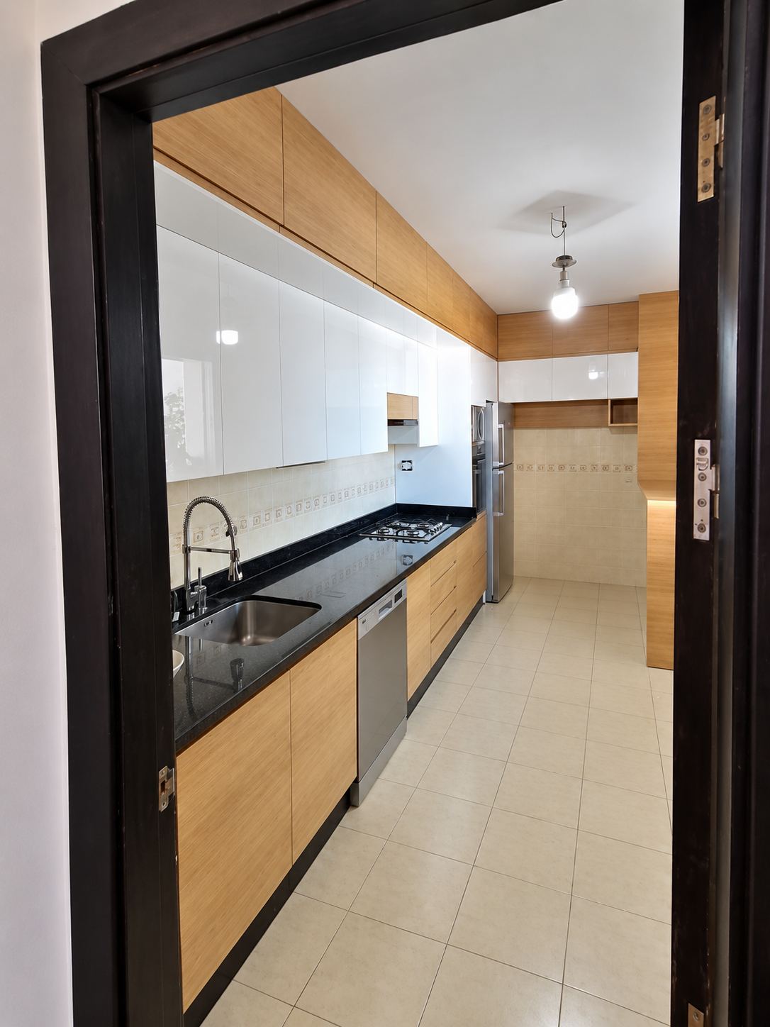 Cuisine en couloir vue depuis l'encadrement d'une porte en bois foncé : façades claires, plan de travail noir et électroménager encastré.