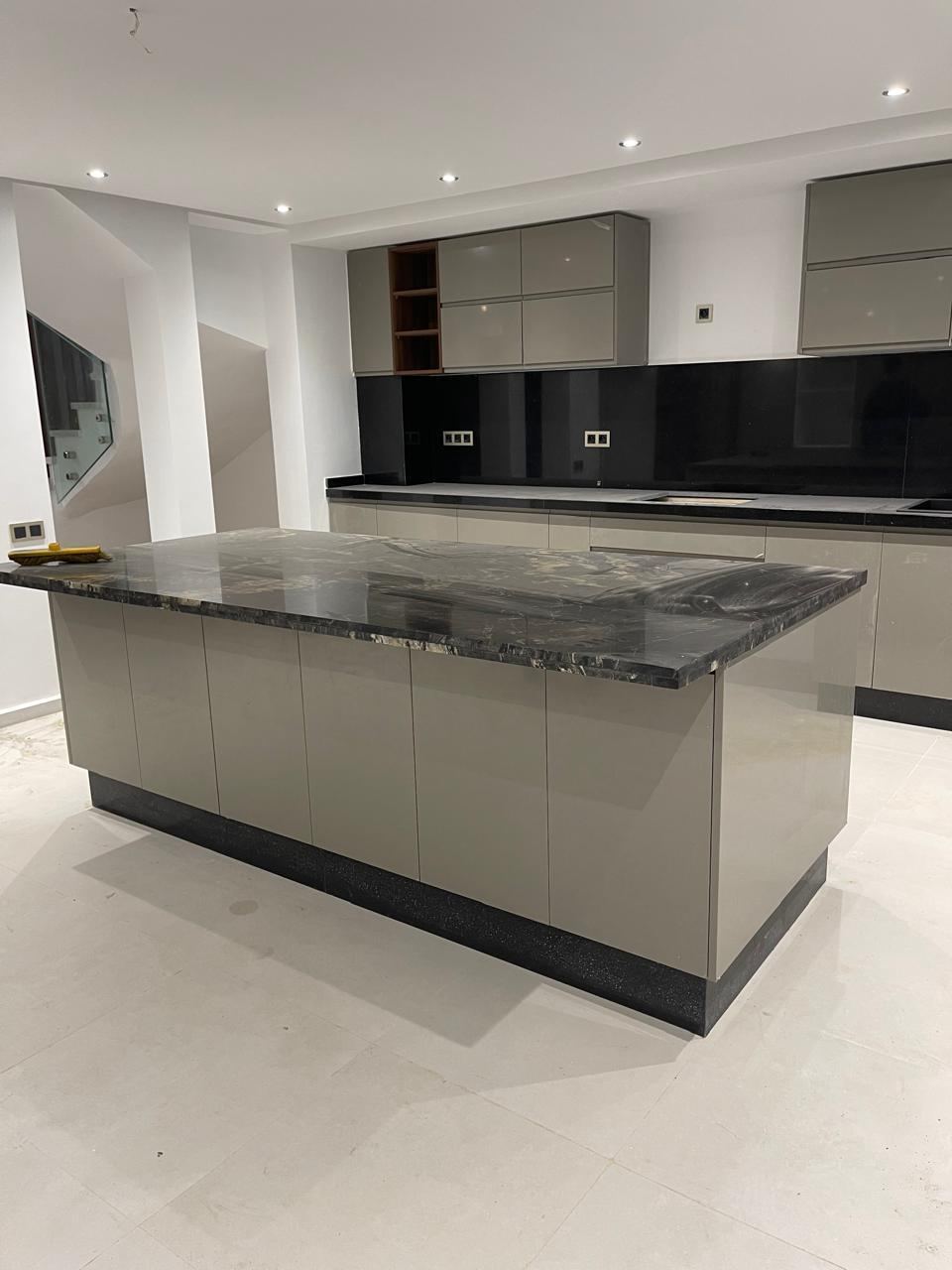 Îlot de cuisine gris clair au plan de travail en granit sombre, face à des meubles muraux gris et une crédence en verre noir.