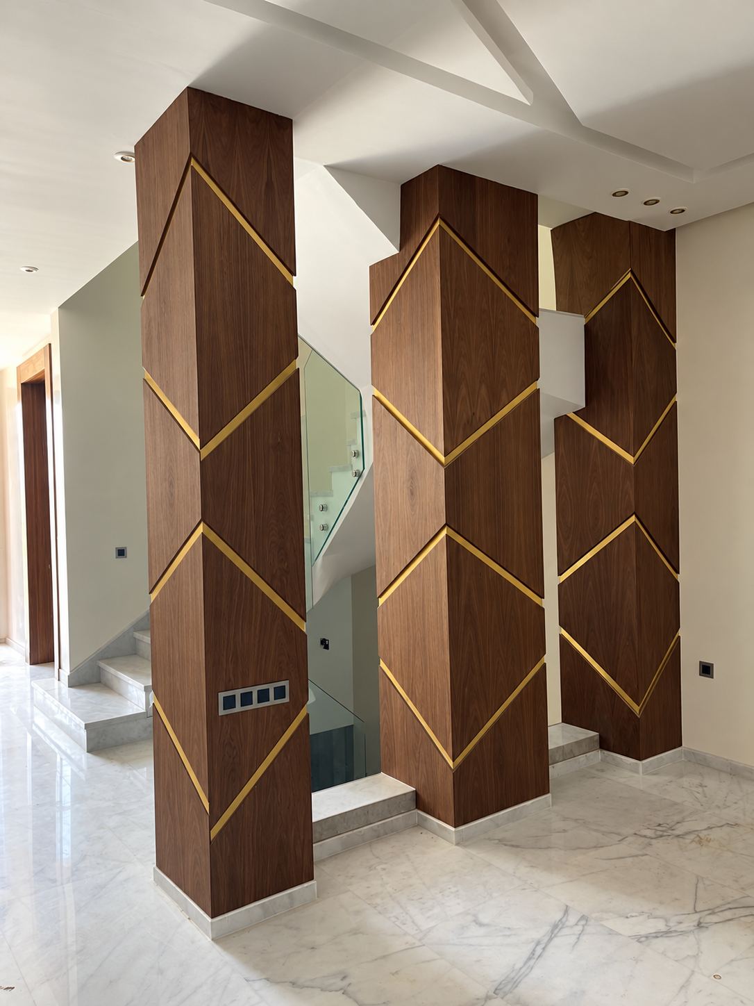 Deux colonnes habillées de panneaux en bois foncé à motif chevron souligné de filets dorés, dans un hall en marbre.