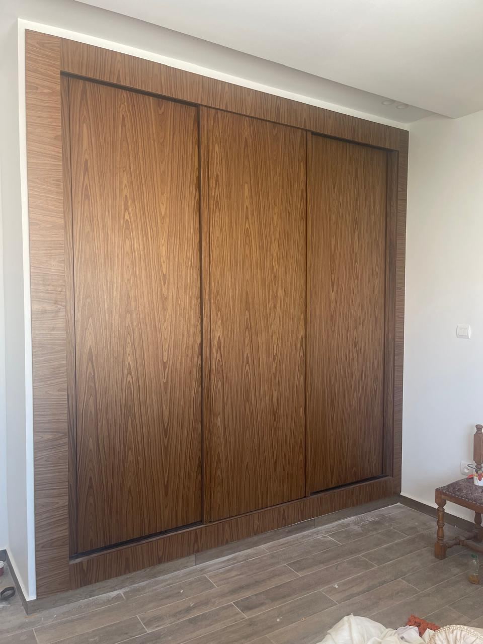 Placard à trois portes coulissantes en bois foncé, encadré d'un caisson lumineux, dans une chambre au sol en bois clair.