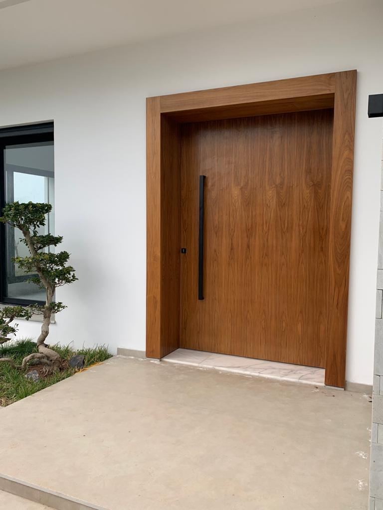 Porte d'entrée pivotante en bois foncé, pleine hauteur, avec longue poignée noire verticale, donnant sur un jardin.