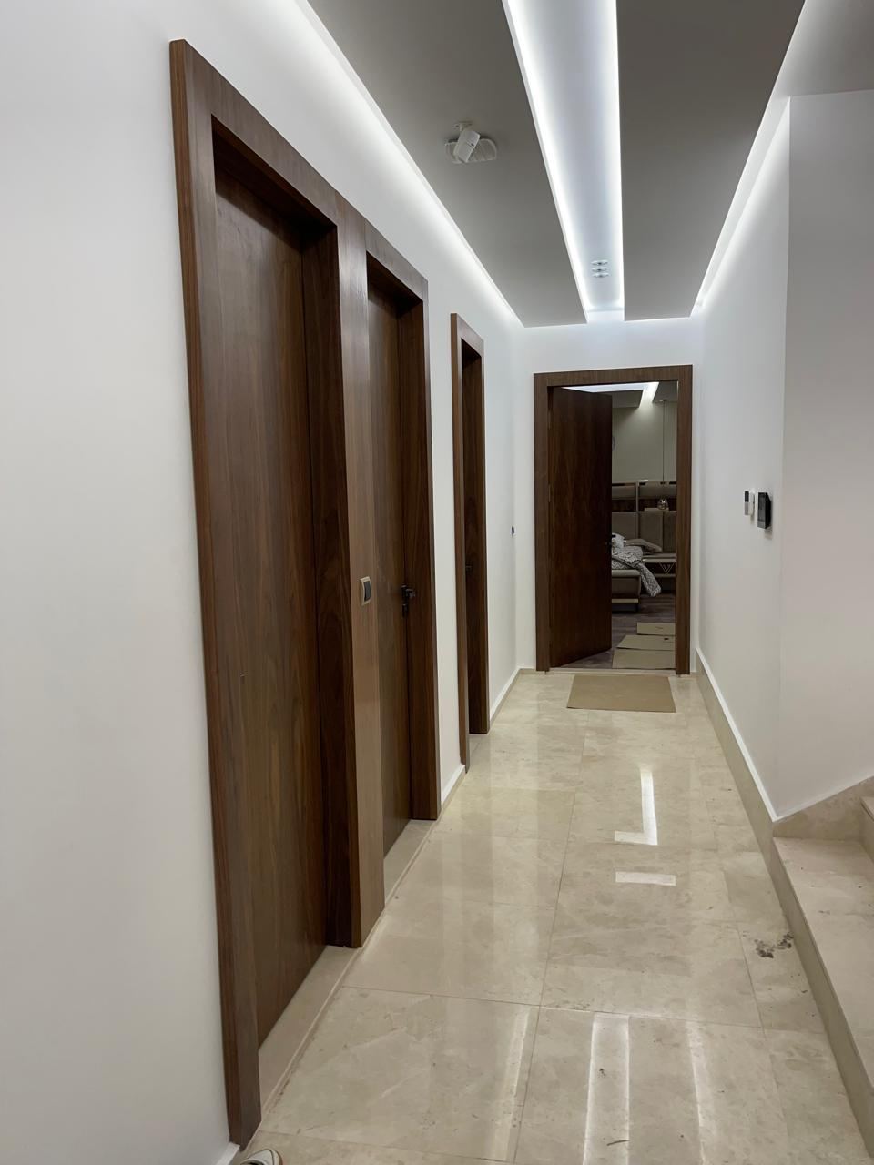 Couloir desservant plusieurs portes intérieures en bois foncé, murs blancs, corniche lumineuse et sol en marbre poli.