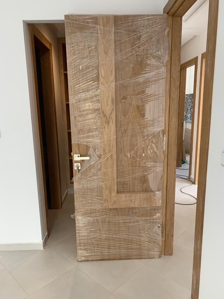 Porte en bois clair à motif géométrique, encore protégée par son film de chantier, posée dans son encadrement.