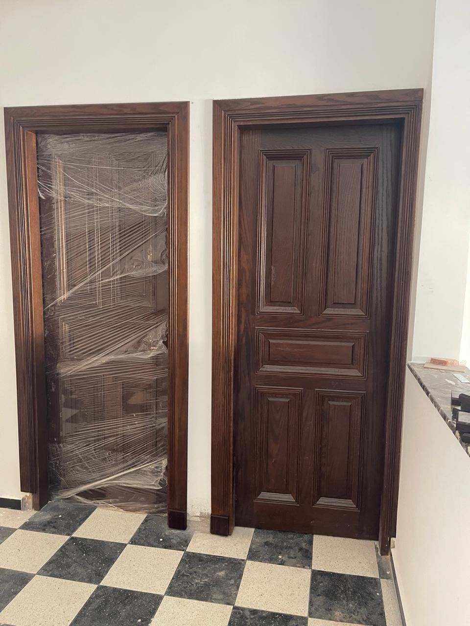 Deux portes à panneaux moulurés en bois foncé, l'une encore sous film de protection, sur un sol en damier.
