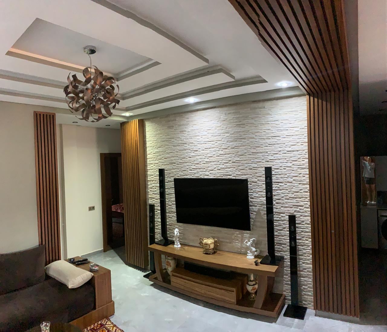 Séjour avec meuble TV sur mesure en bois, mur en pierre claire et faux plafond à tasseaux de bois sur un côté.