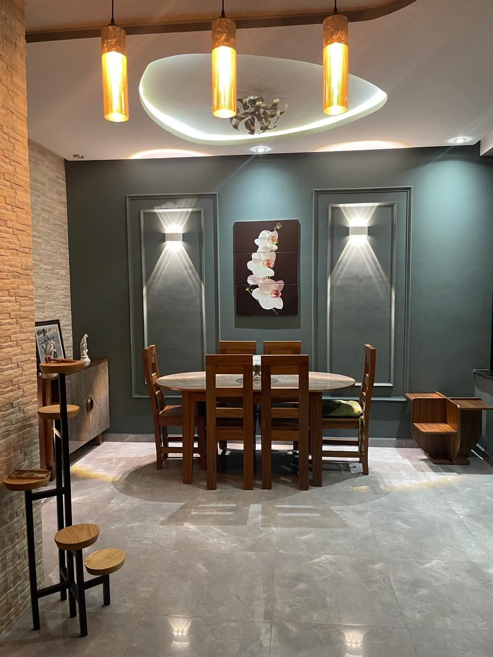 Coin repas avec table et chaises en bois, mur sombre, panneaux muraux éclairés et trois suspensions dorées.